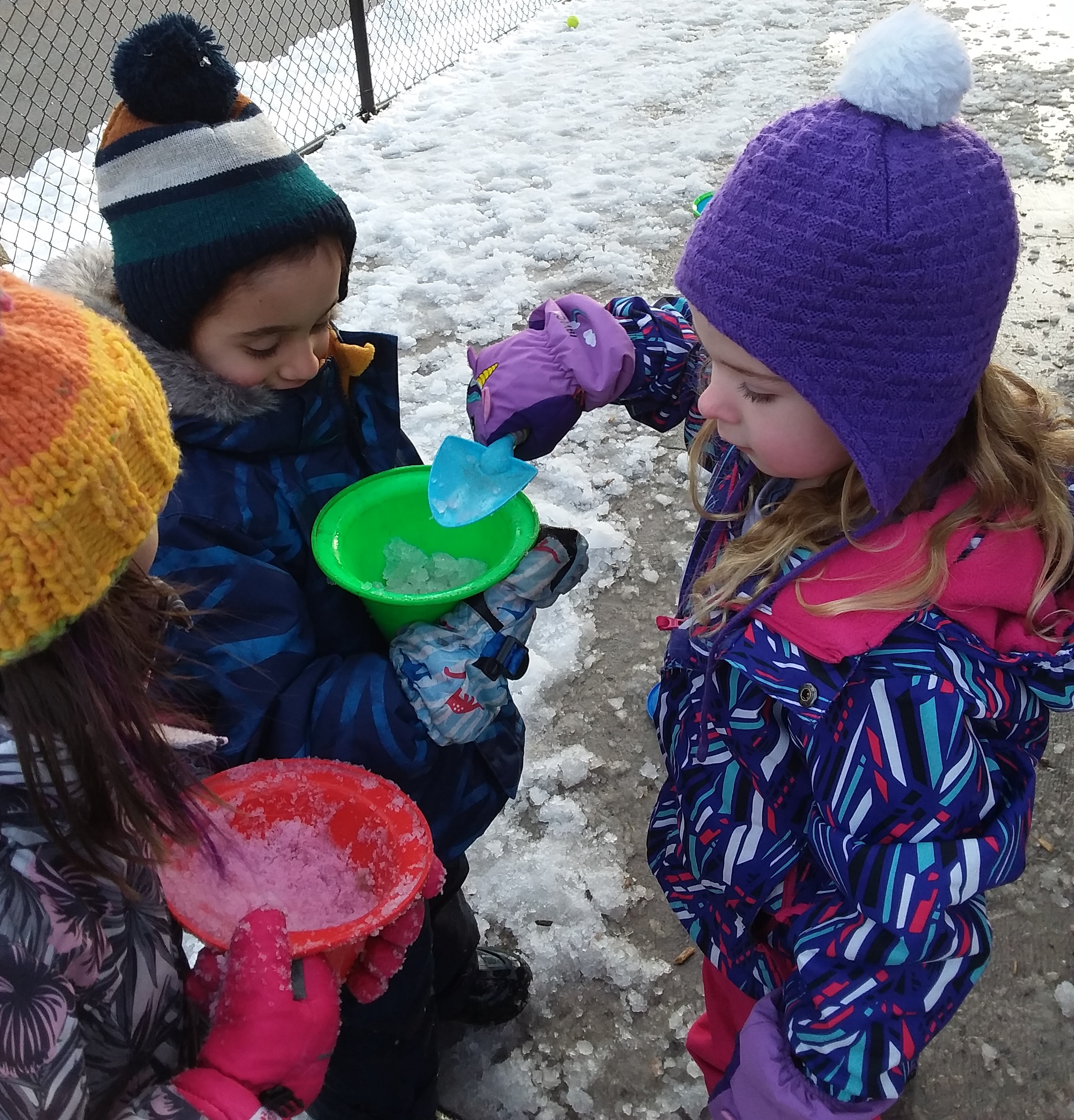 A group of young children in a PLASP after school program play with coloured snow using shovels and buckets in a school play ground