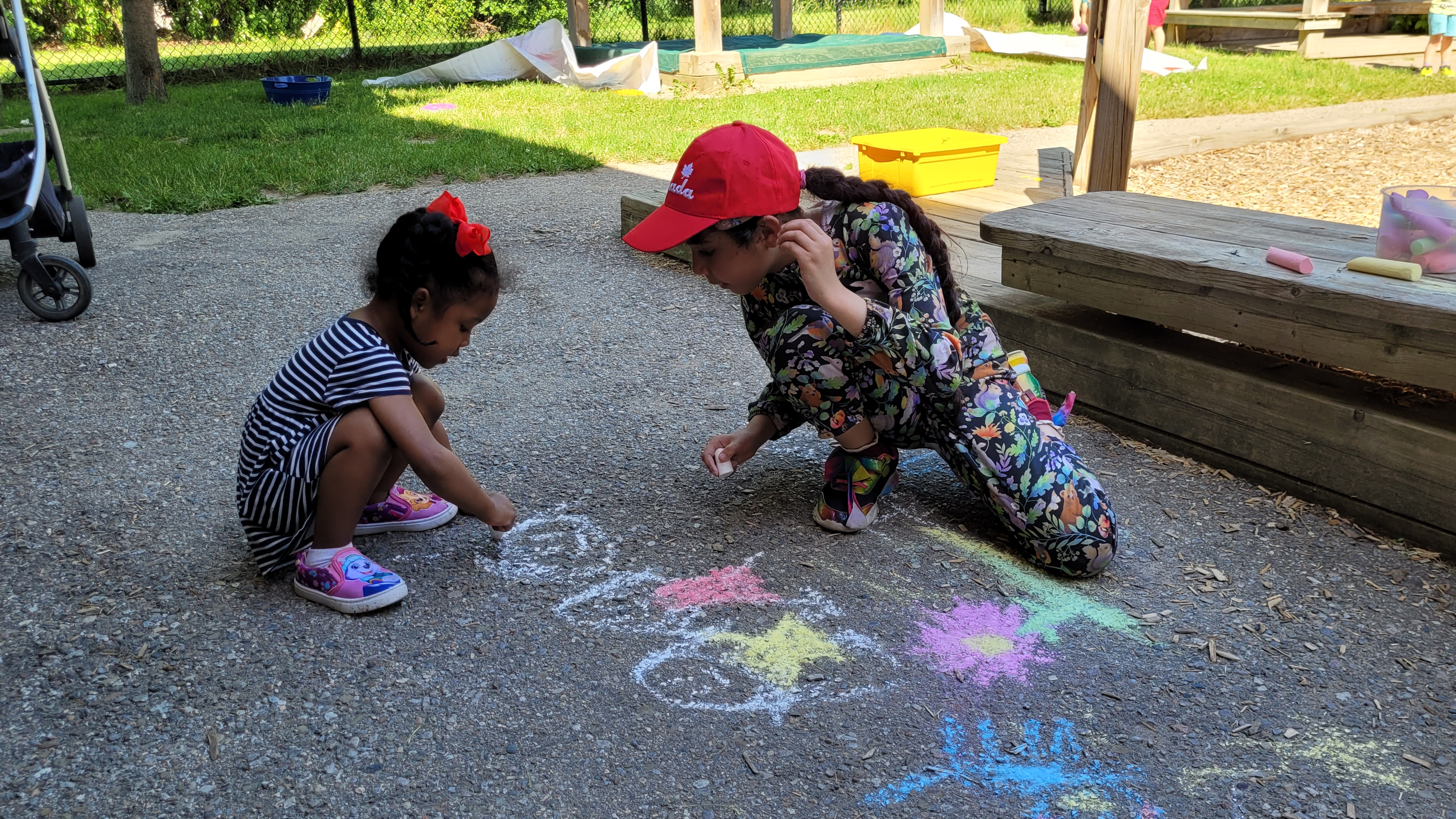 Two children crouch down in a playground to draw on the ground with sidewalk chalk