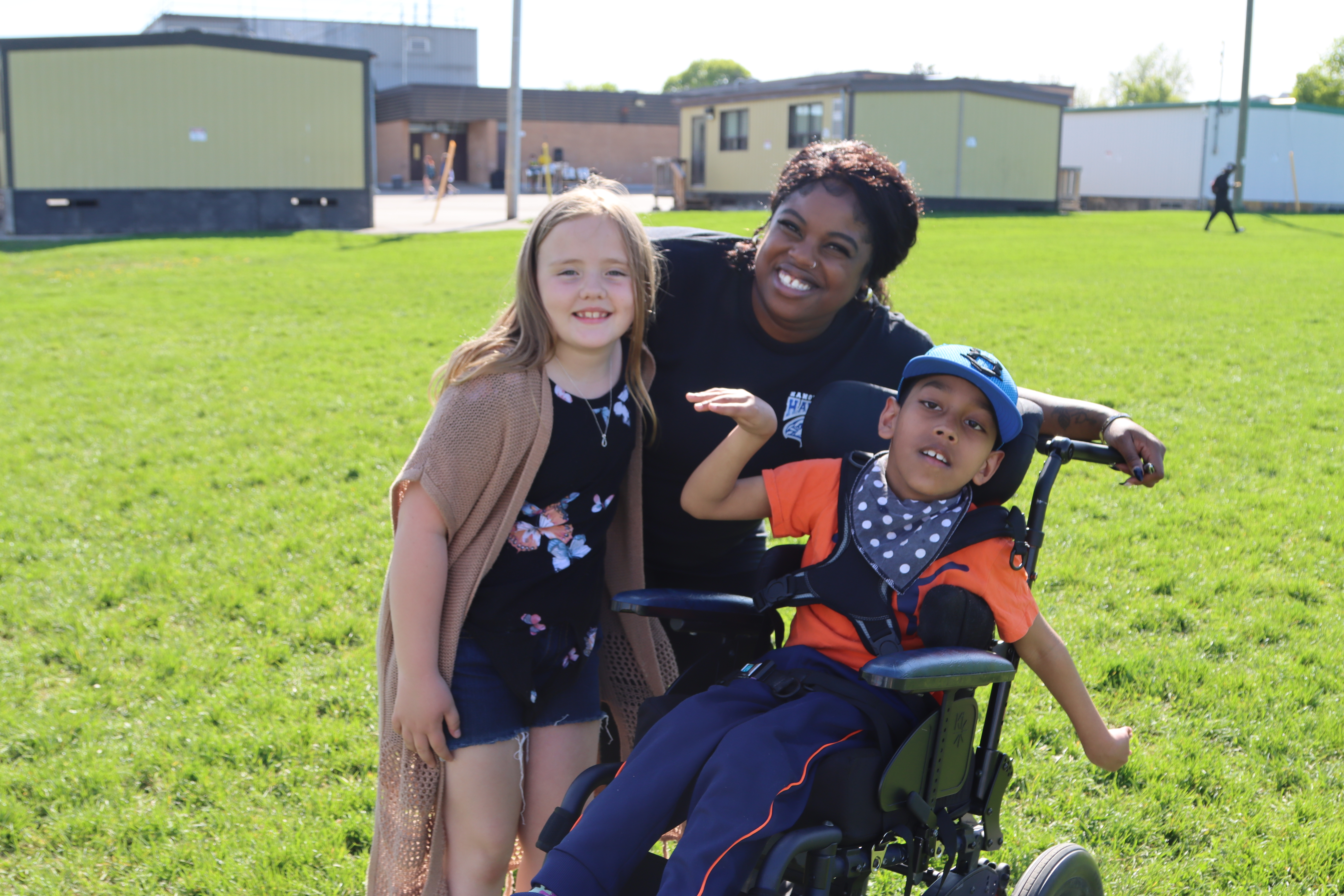 A PLASP educator stands in between two children on a school field, with an arm around each one, and everyone is smiling 