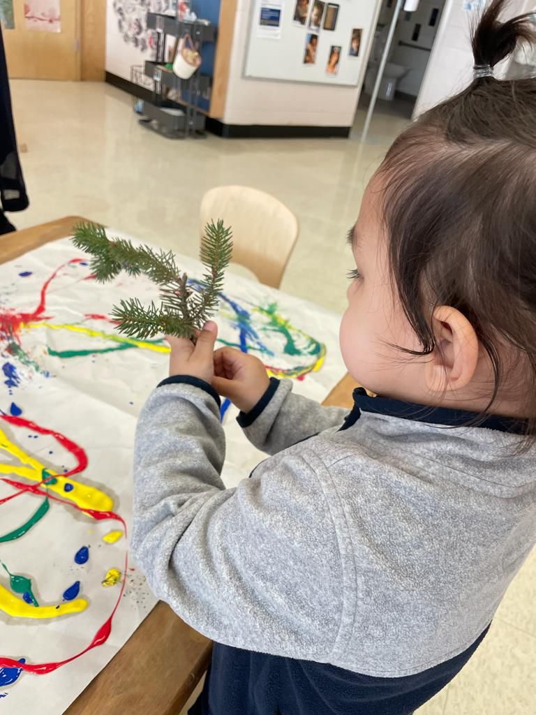 A young child holds a small pine tree branch and stands in front of a table where a piece of white paper with yellow, blue, red, and green swirls painted on it