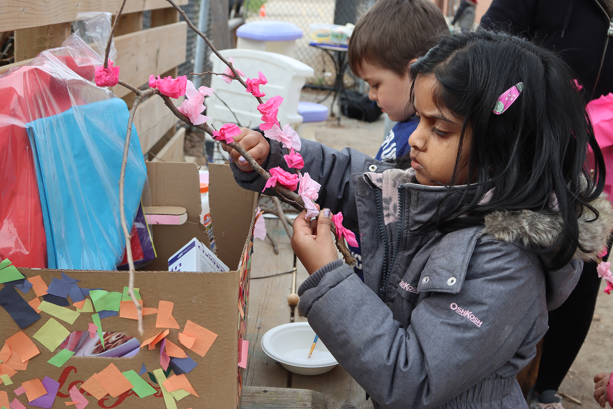 Two kindergarten children make cherry blossoms outside using sticks and pink tissue paper