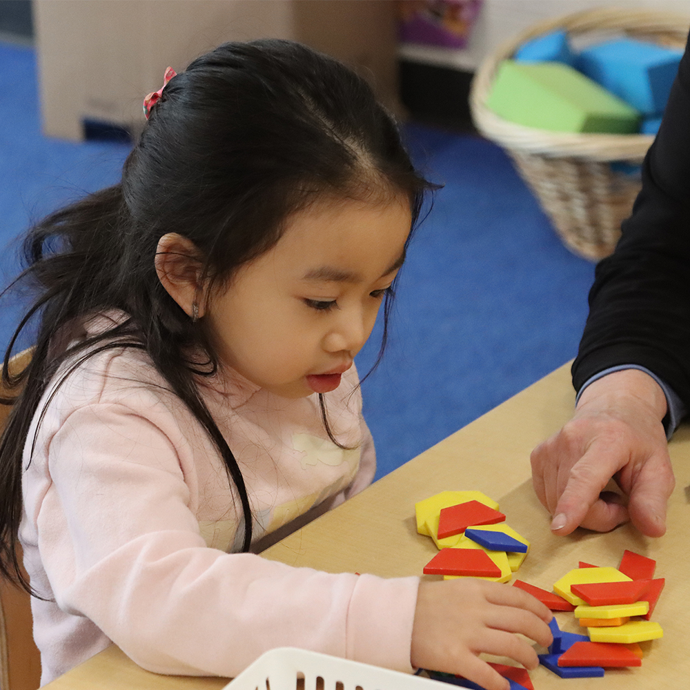 A young child sits at a table in a PLASP program exploring yellow, red, and blue shapes