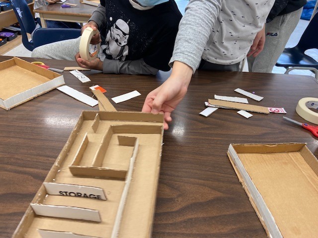 A picture of children’s hands creating a cardboard maze using tape, scissors, and strips of cut up cardboard