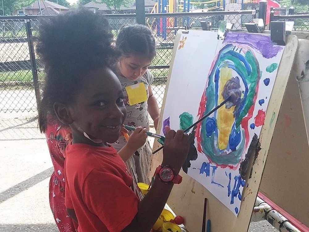 A young girl stands in front of an easel holding a paintbrush outside at a PLASP program