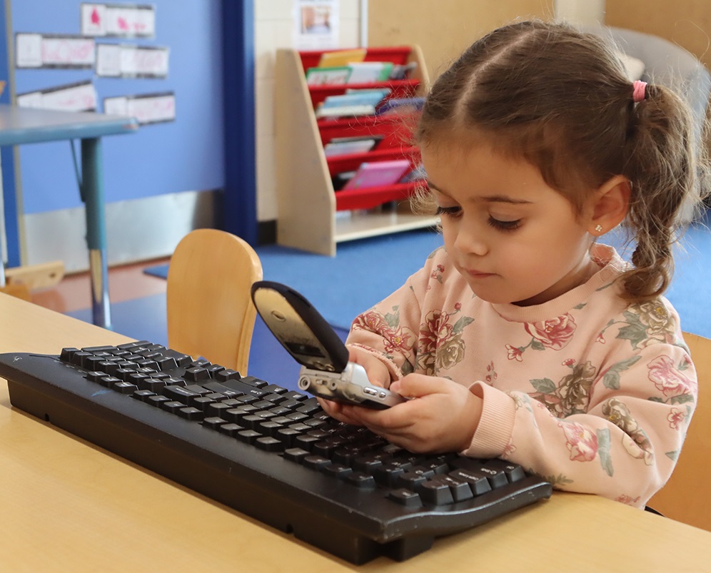 A young girl in a PLASP program sits at a table in a classroom at a computer keyboard and uses a flip phone