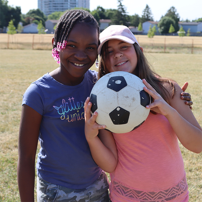 Two smiling girls on a playground in the sunshine, one is holding a soccer ball